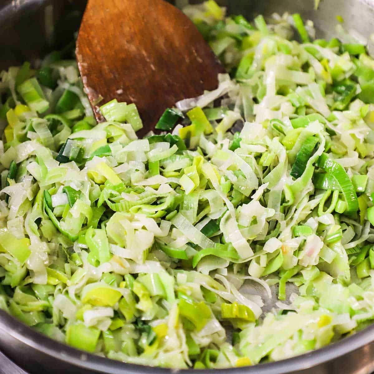 Chopped leeks and onions being sautéd in a large skillet with a wooden spatula stirring the veggies.