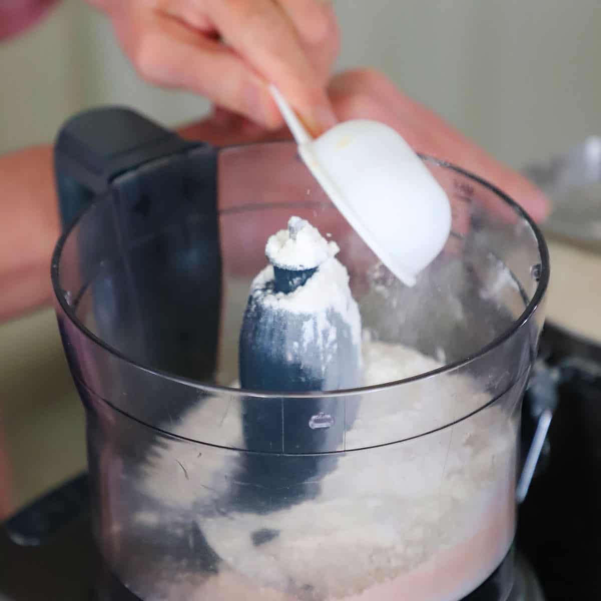 A person using a white measuring cup to transfer flour into a food processor.