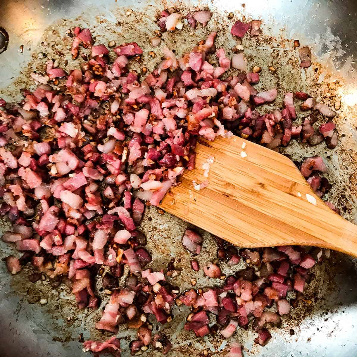 Cubed pancetta being sautéed in a large skillet being stirred with a wooden spatula.