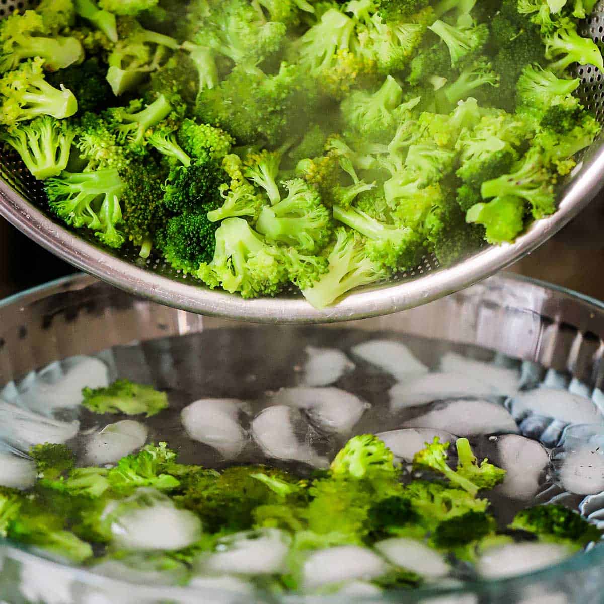 Broccoli florets that have been blanched in boiling water being transferred to an ice batch in a glass bowl.