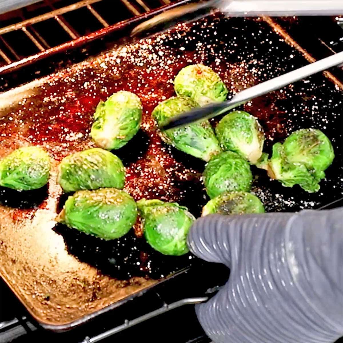 A person using oven mitts to remove a baking sheet from the oven that has roasted Brussels sprouts on it.