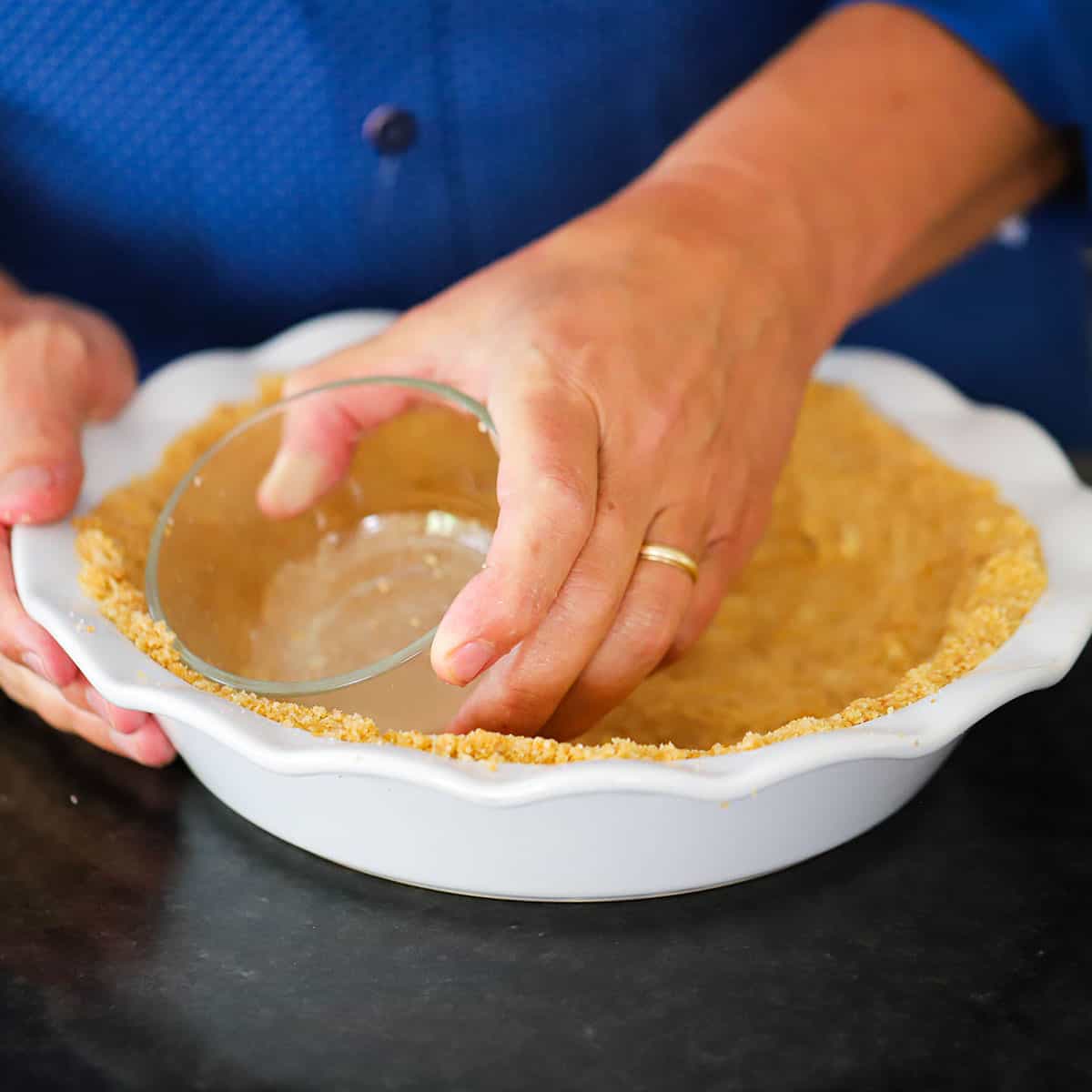 A person using a small glass bowl to press a graham cracker crust into the sides of a white pie dish.