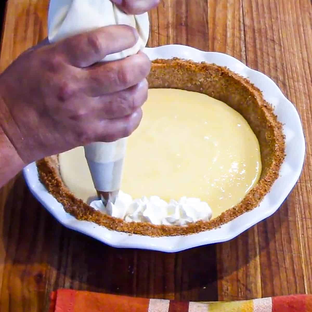 A person piping whipped cream from a pastry bag onto the top perimeter of an easy key lime pie.
