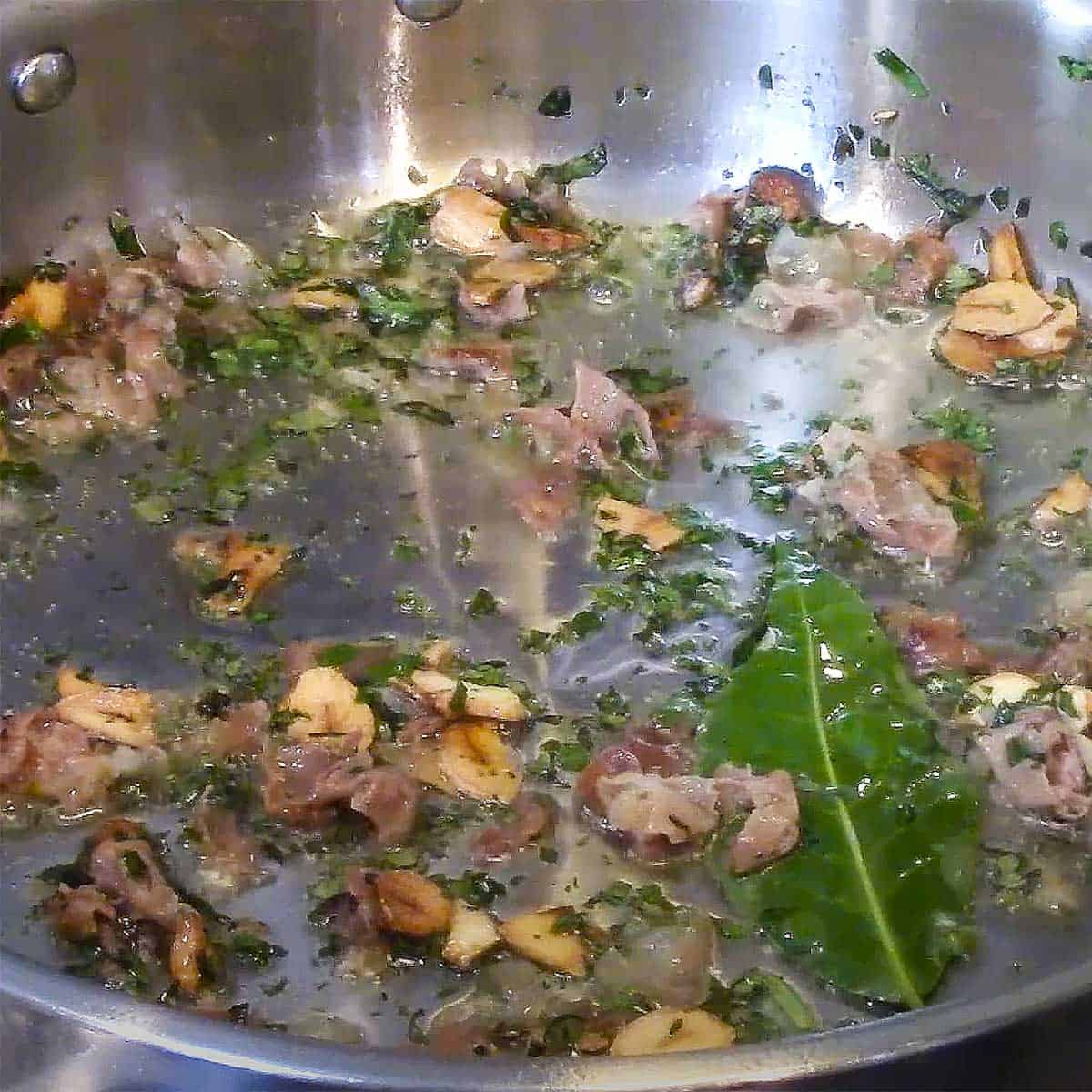 A skillet filled with simmering garlic, prosciutto, herbs, and a bay leaf.
