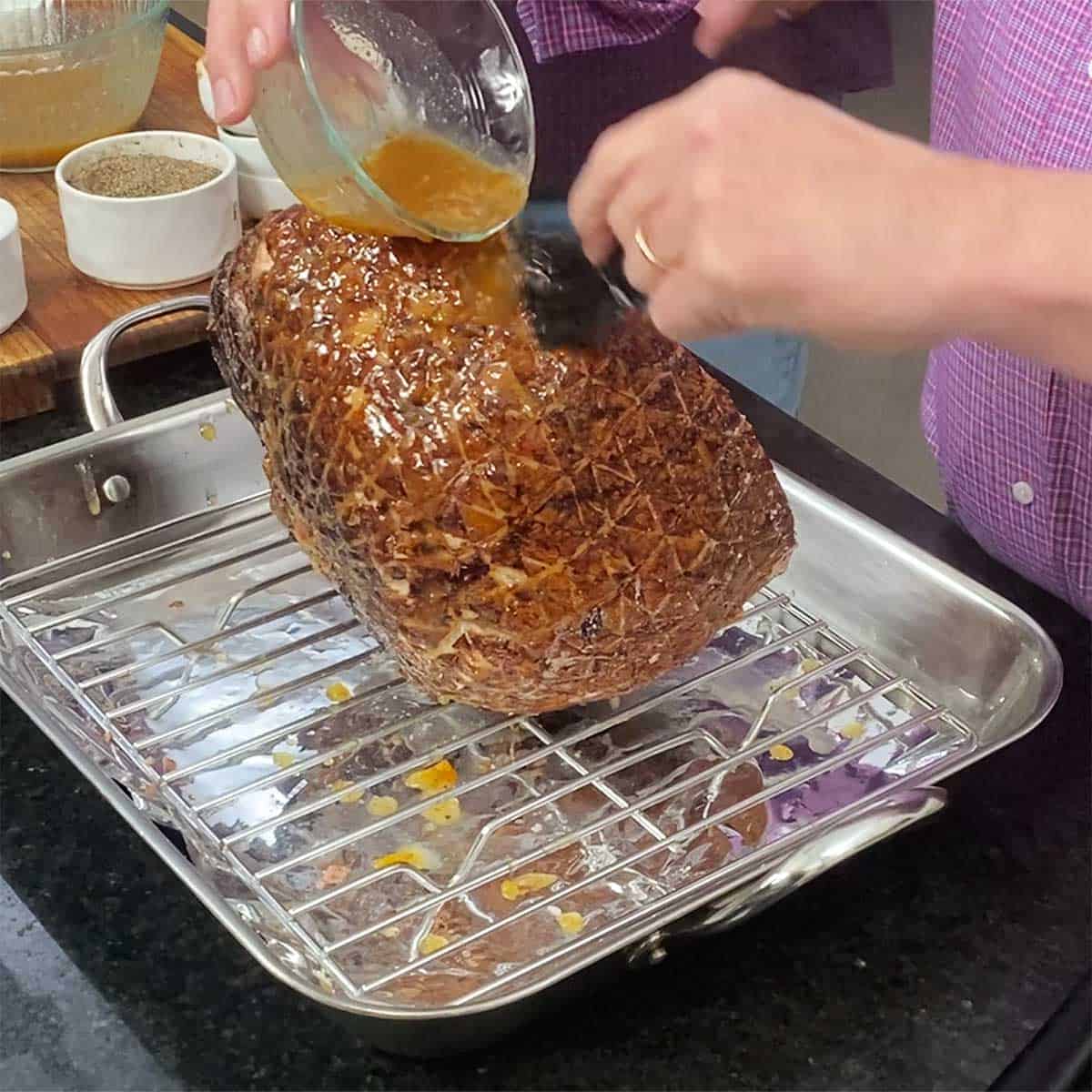 A person brushing a bourbon orange glaze over the exterior of a smoked ham that is resting on a roasting rack in a roasting pan.