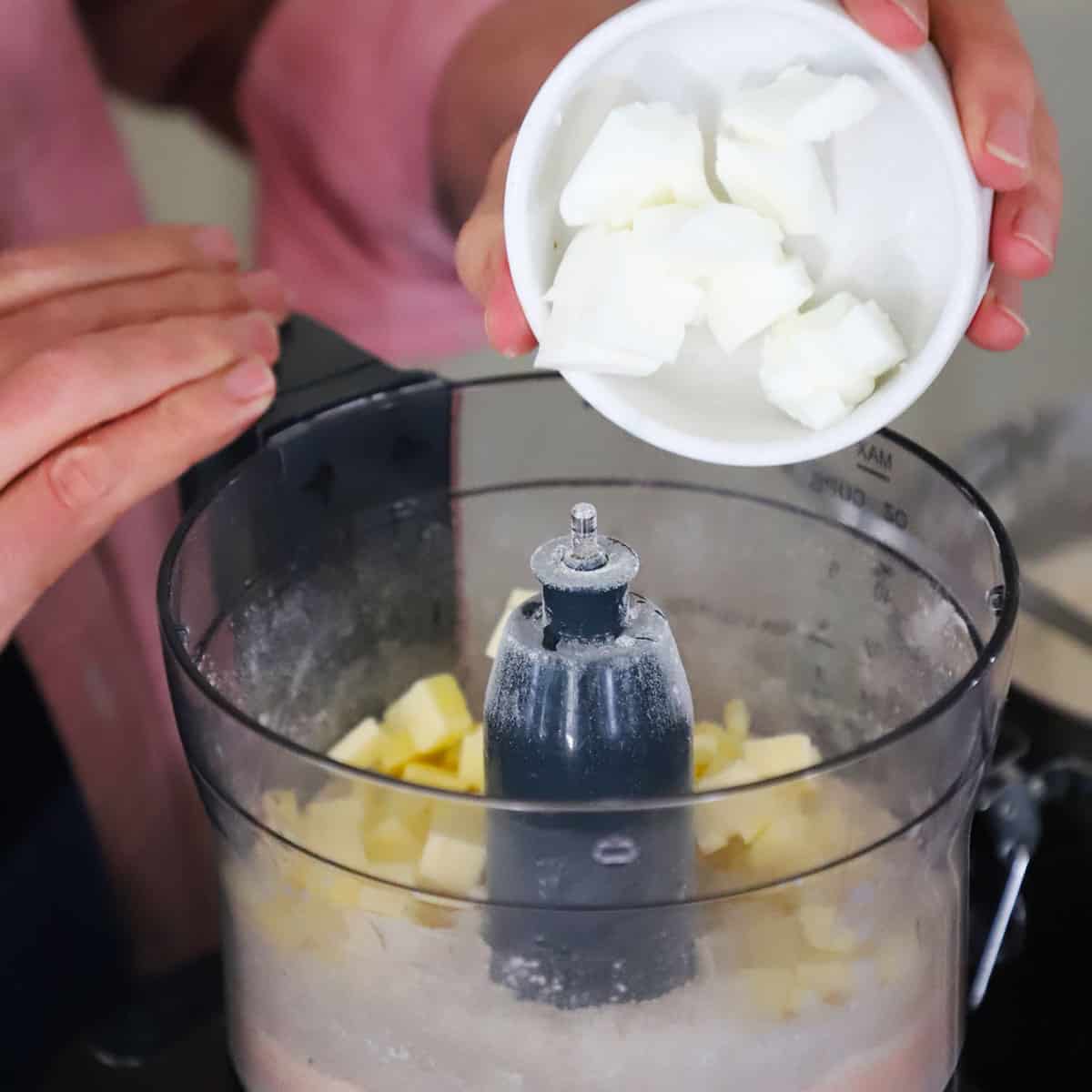 A person transferring chilled cubed shortening and butter into the base of a food processor that already contains flour and salt.