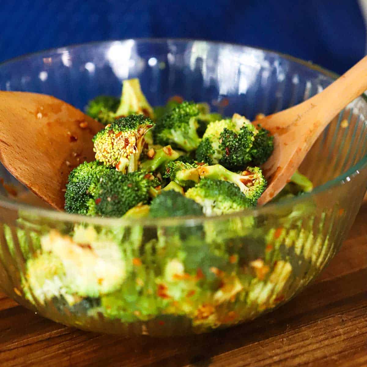 A person using two wooden spatulas to toss broccoli florets in a glass bowl with a chili sauce and soy sauce mixture.