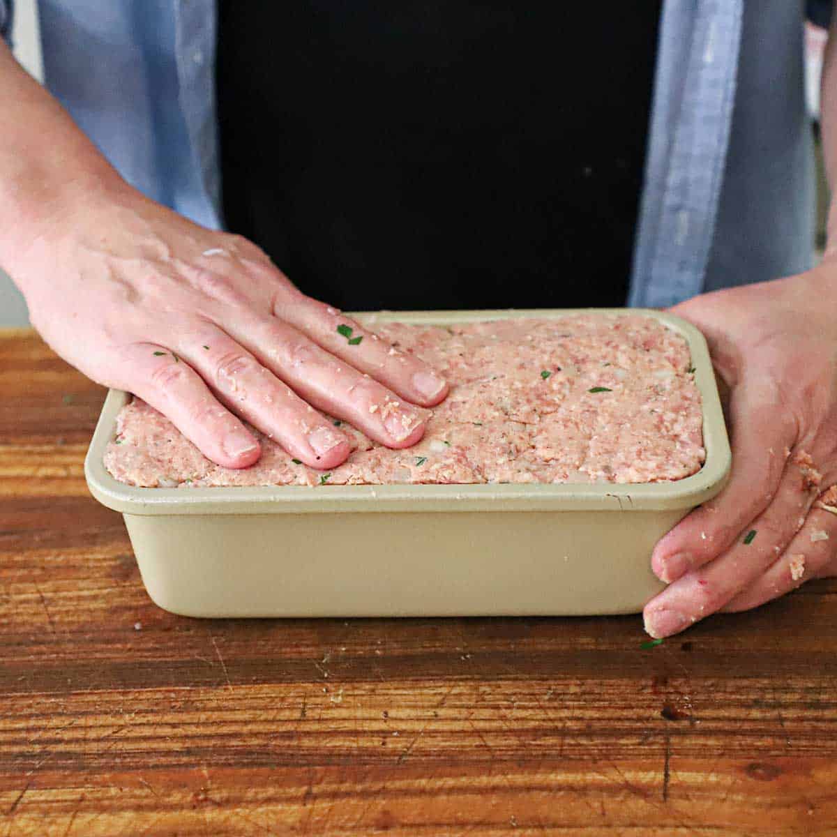 A person pressing compacting a ham mixture for ham loaf into a metal loaf pan on a wooden cutting board.