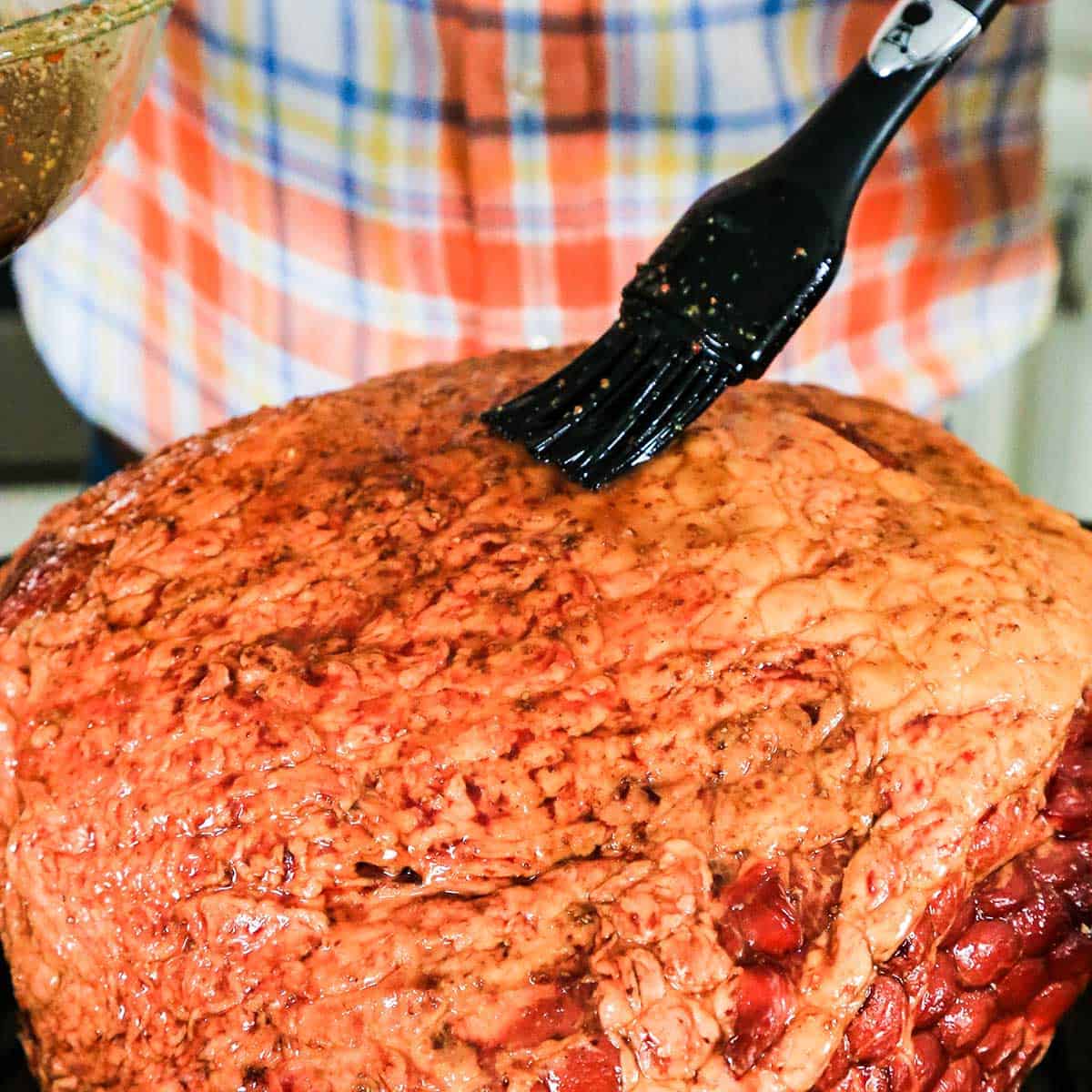 A person using a pastry brush to apply a honey bourbon glaze over the top of a roasted and smoked Berkshire ham that has been scored with a knife.