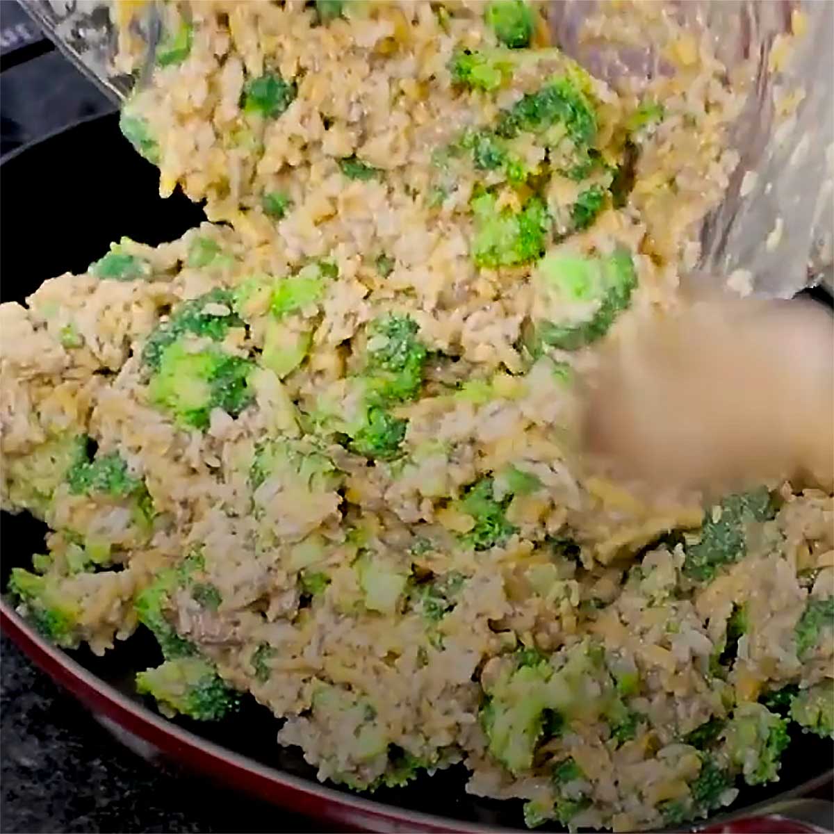 A person transferring a mixture of cream of mushroom soup, broccoli, cheddar, and rice into an oval baking dish.