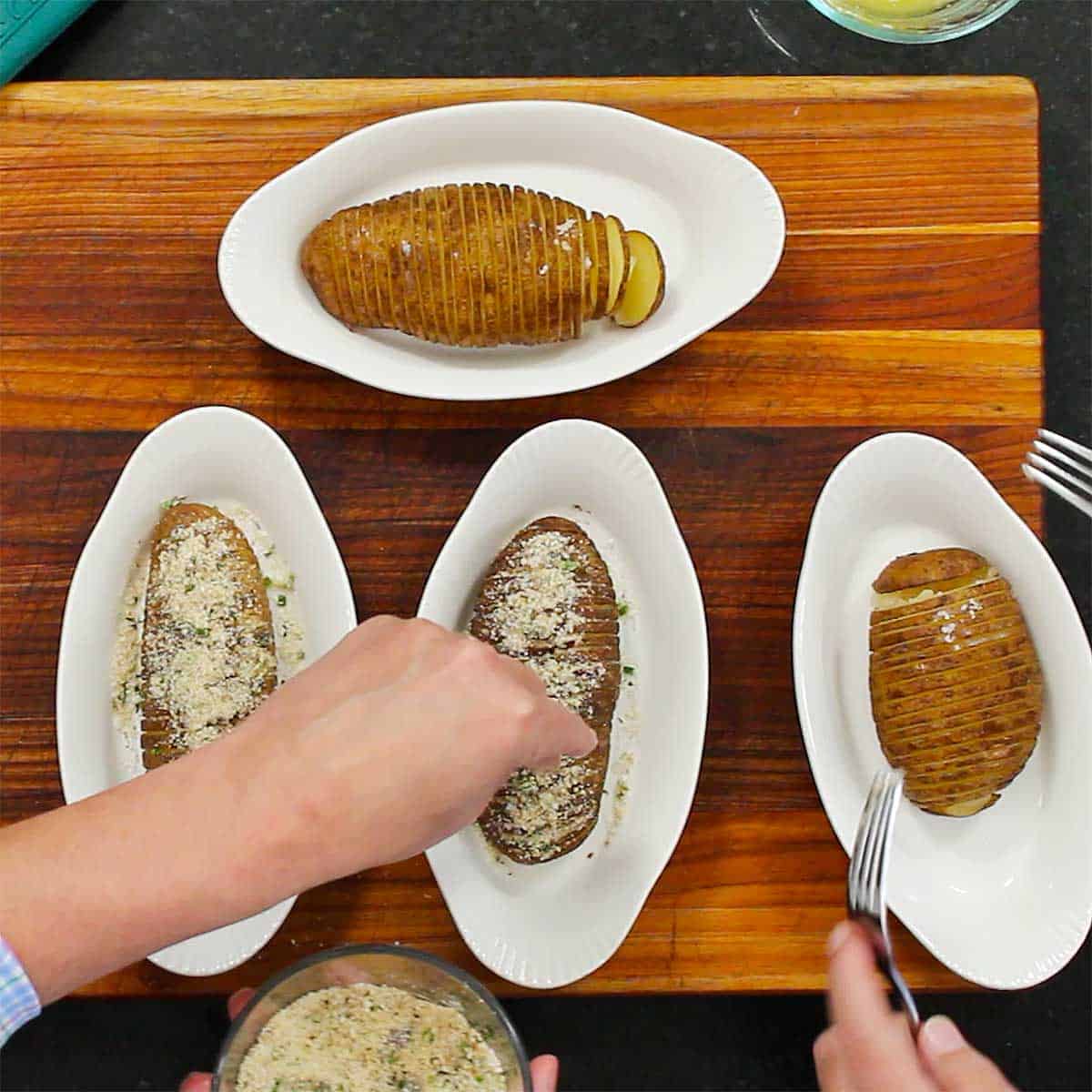 A person sprinkling breadcrumbs and grated Parmesan cheese on the tops of hasselback potatoes that are each in their own individual baking dish.