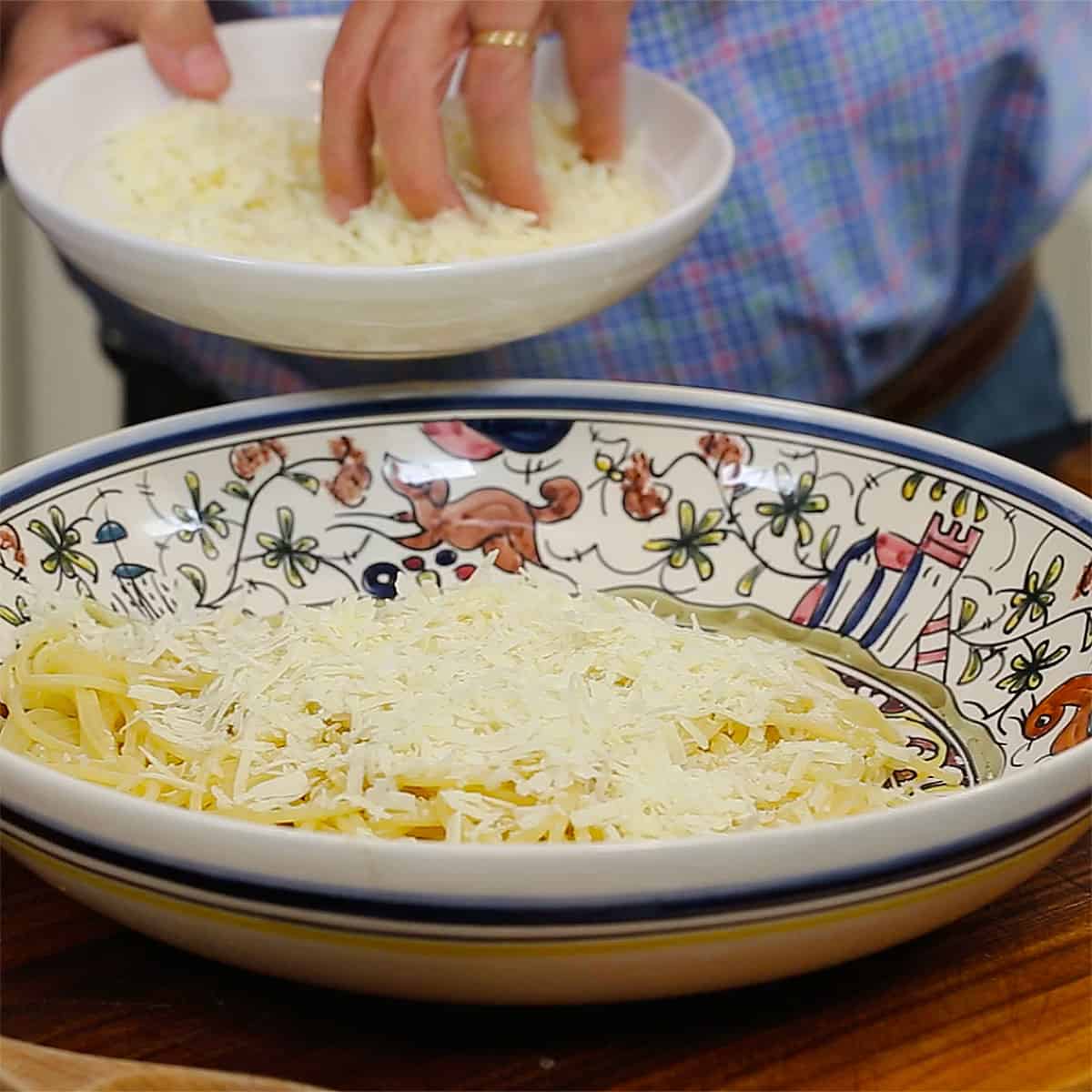A person placing grated cheese into a large pasta bowl filled with cooked pasta.