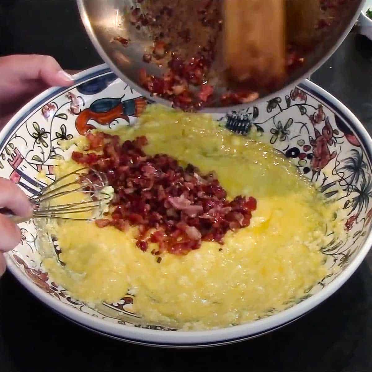 A person transferring cooked cubed pancetta into a shallow pasta bowl filled with an egg and cheese mixture.