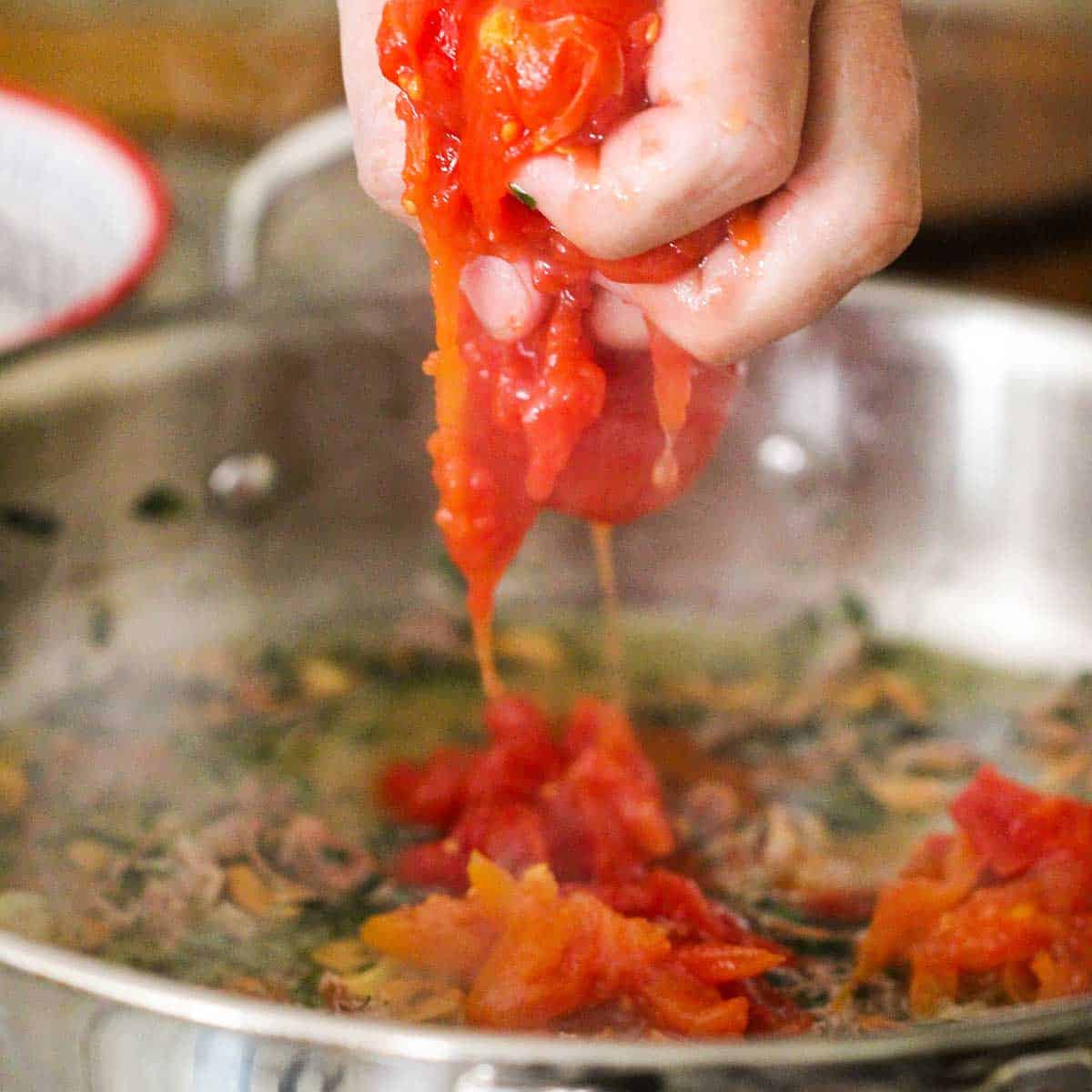 A person squeezing canned tomatoes into a skillet filled with simmering garlic, prosciutto, and herbs.