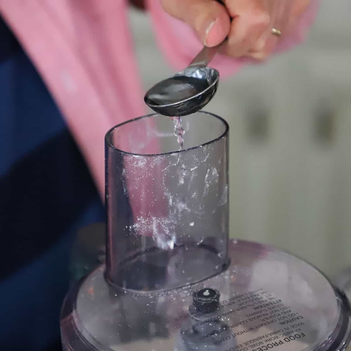 A person using a tablespoon to add water into the tube of a food processor filled with pie dough.
