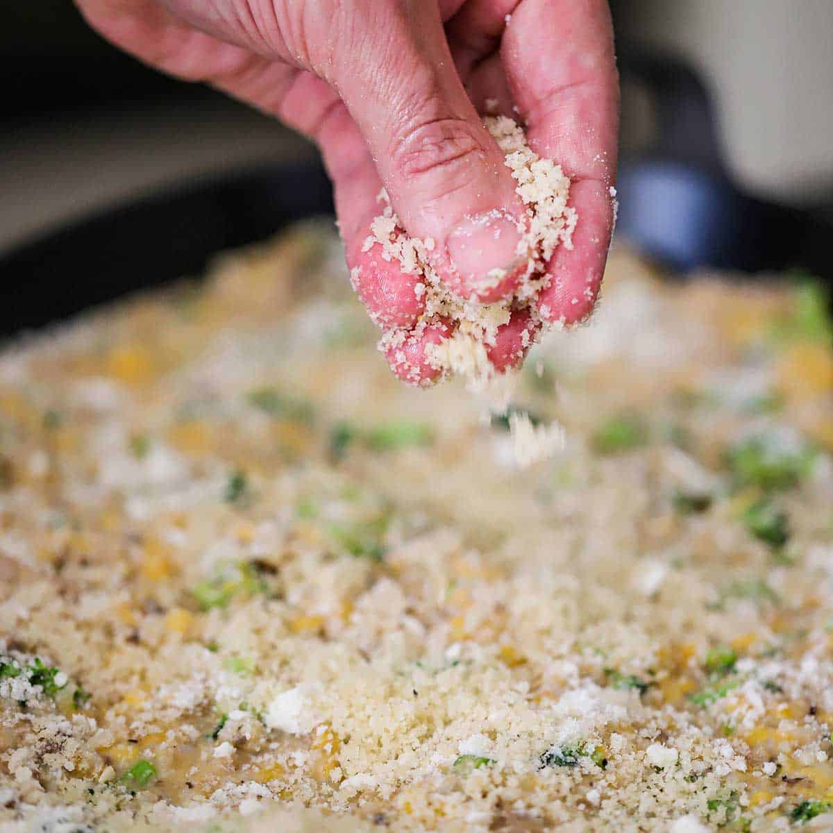 A person sprinkling breadcrumbs with grated Parmesan cheese over the top of an uncooked broccoli and cheddar casserole.