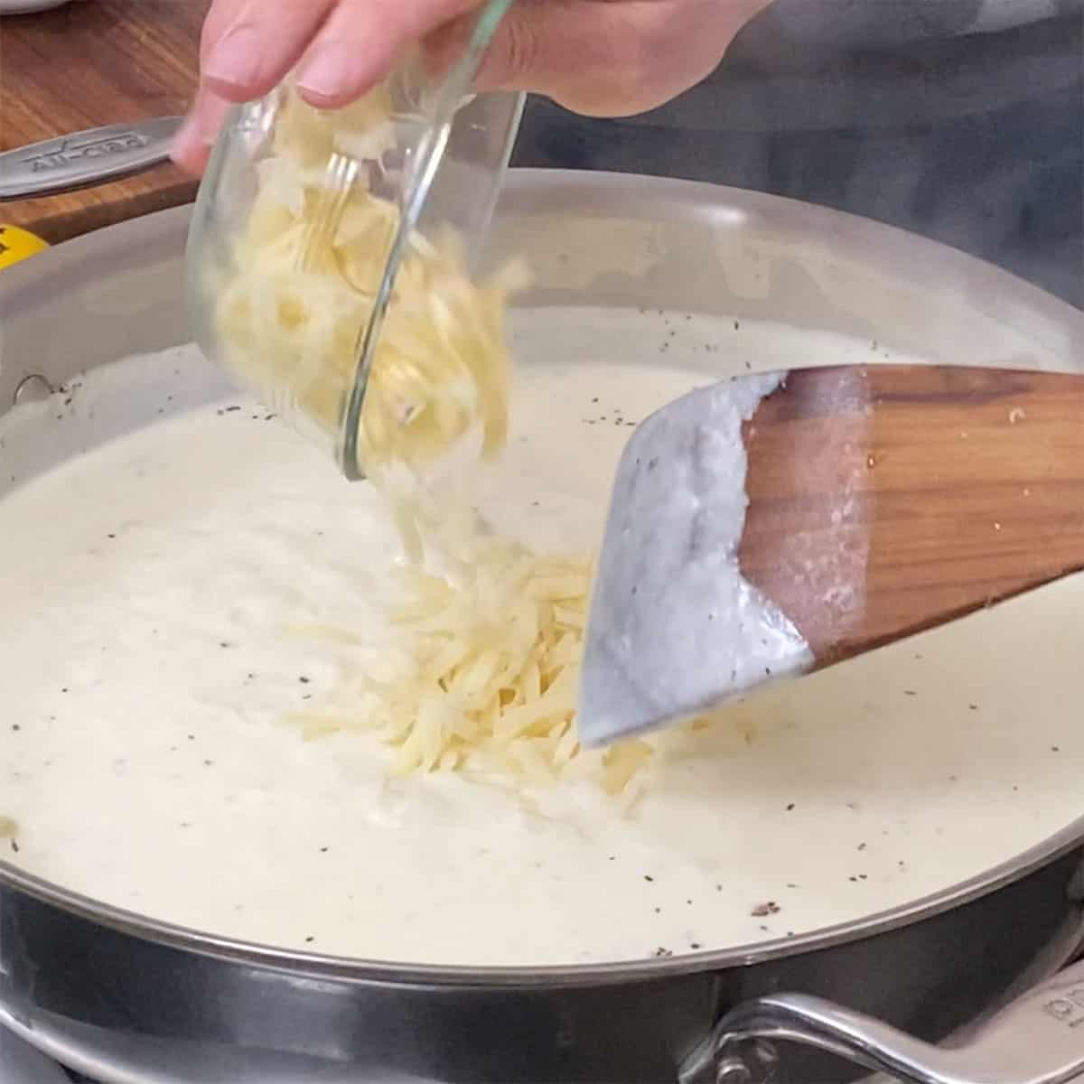 A person adding shredded cheese into a skillet that is filled with a creamy sauce.