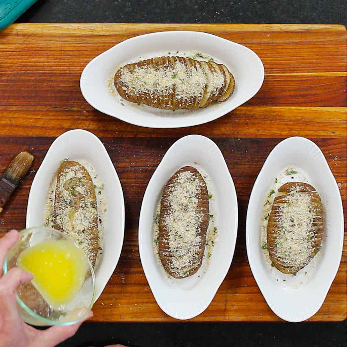 A person drizzling melted butter over the tops of baked hasselback potatoes.