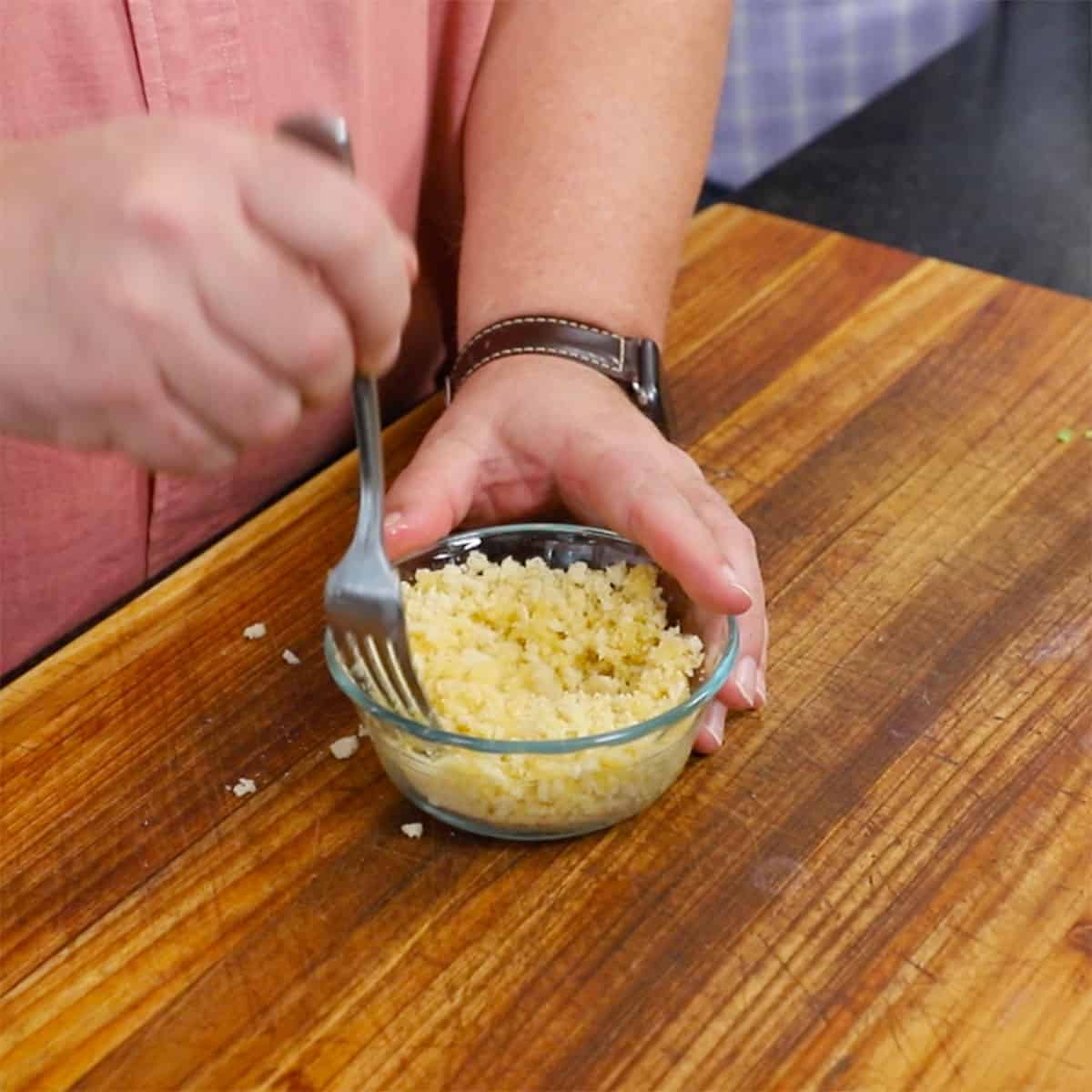 A person using a fork to combine melted butter in a small bowl filled with breadcrumbs.