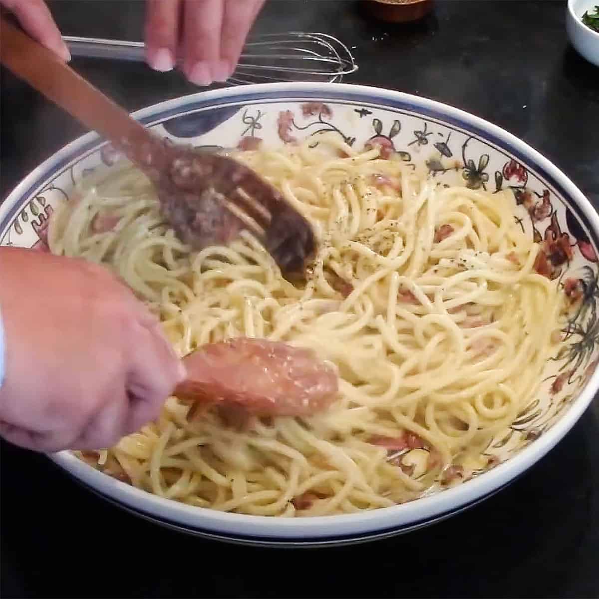 A person using two wooden utensils to toss cooked pasta with an egg and cheese mixture with crispy pancetta.