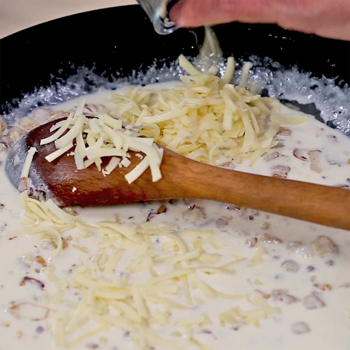 A person adding shredded Gruyere cheese into a skillet of simmering cream and sautéd onions and pancetta.