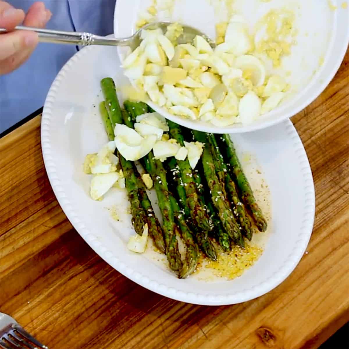 A person transferring chopped eggs in a lemon vinaigrette onto a platter filled with sautéed asparagus.