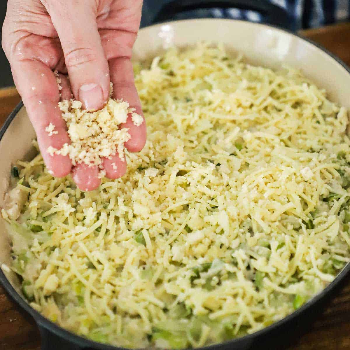 A person sprinkling breadcrumbs over the top of leek gratin in an oval baking dish.