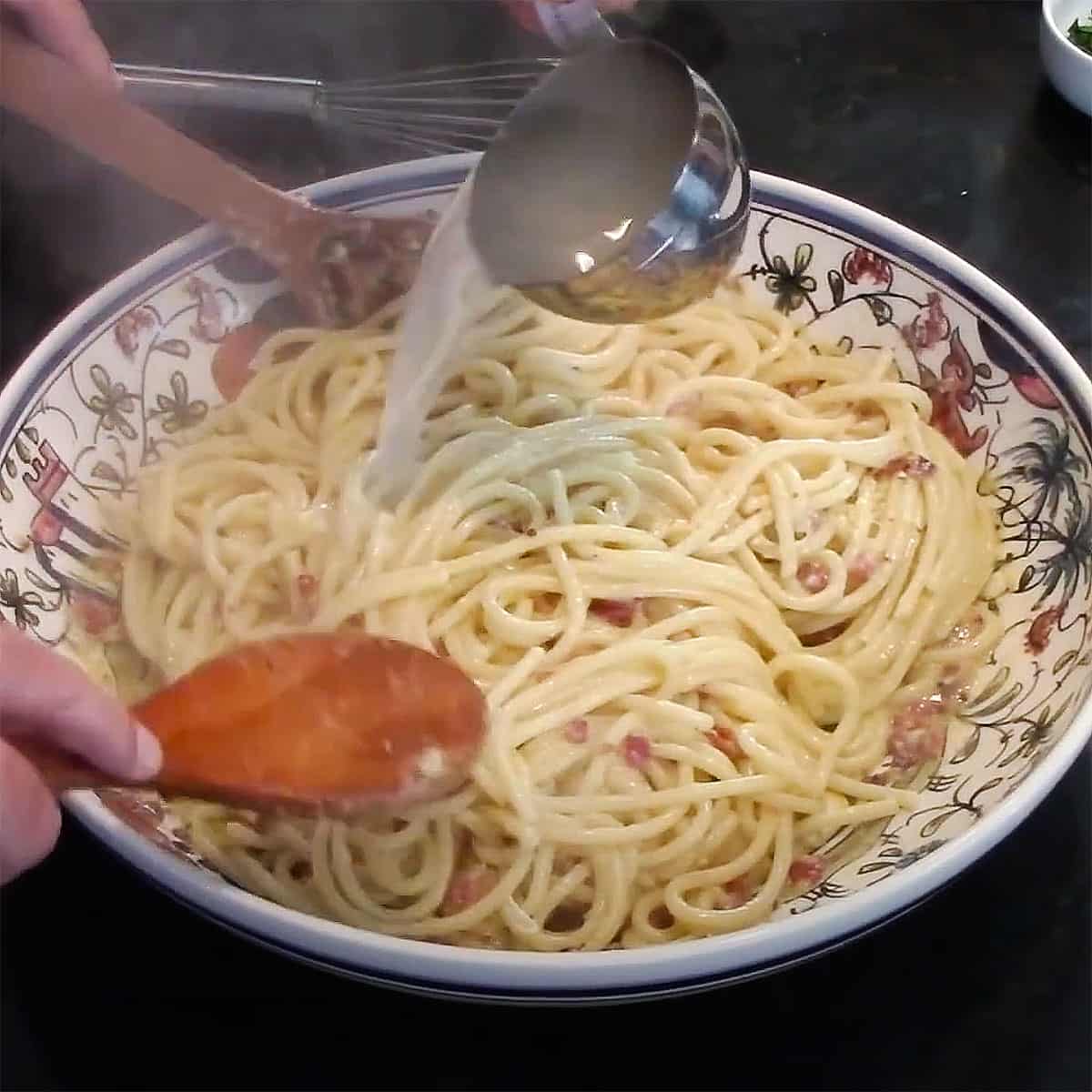 A person adding pasta water from a metal measuring cup into a large pasta bowl filled with pasta carbonara.