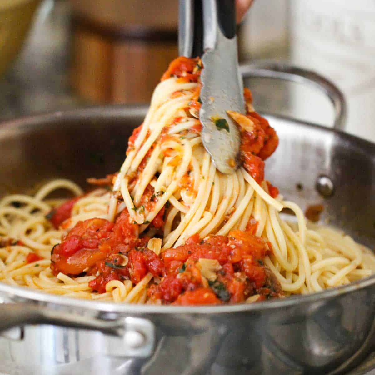 A person using a pair of metal tongs to toss cooked pasta with sautéed tomatoes and herbs.