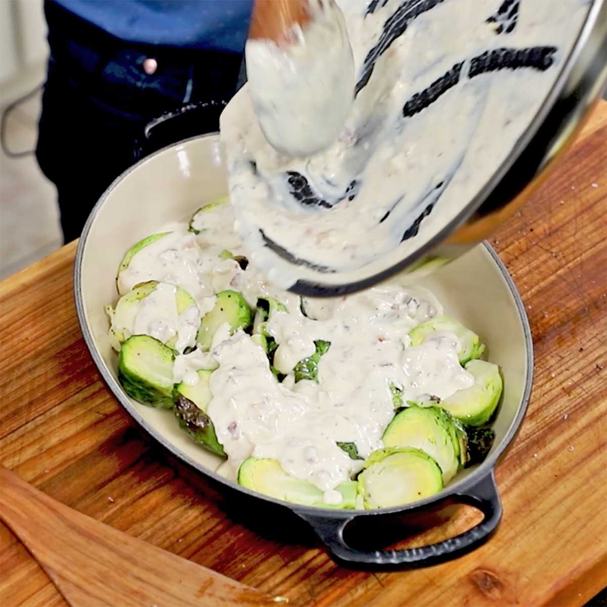 A person transferring a béchamel sauce from a skillet into a baking dish that is filled with roasted Brussels sprouts.