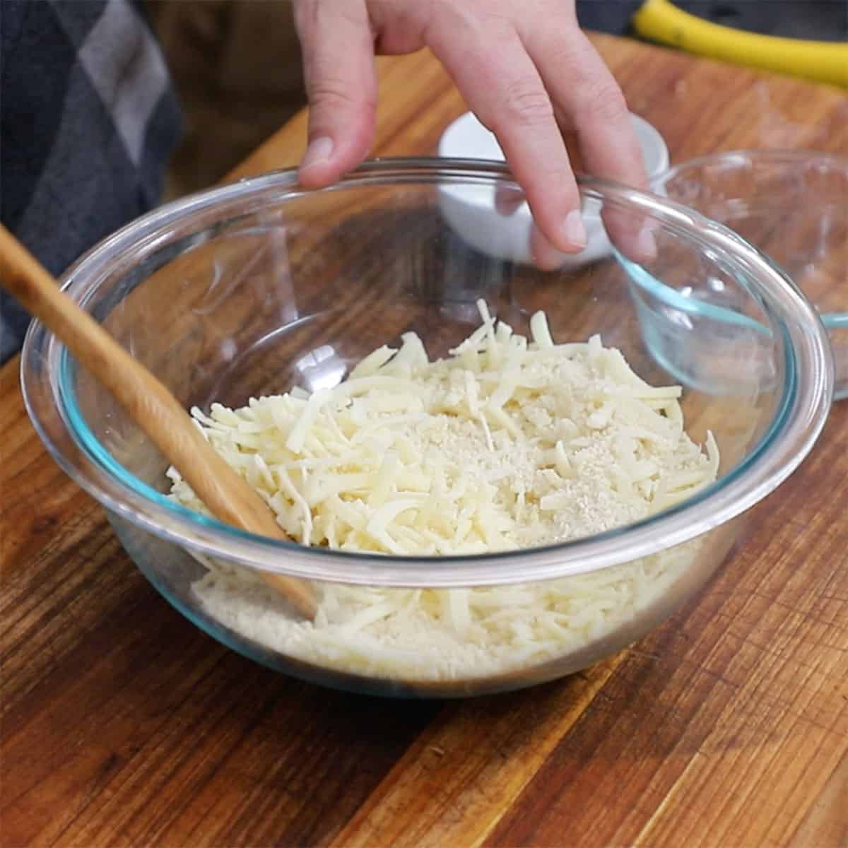 A person combing shredded cheese with breadcrumbs in a glass bowl.