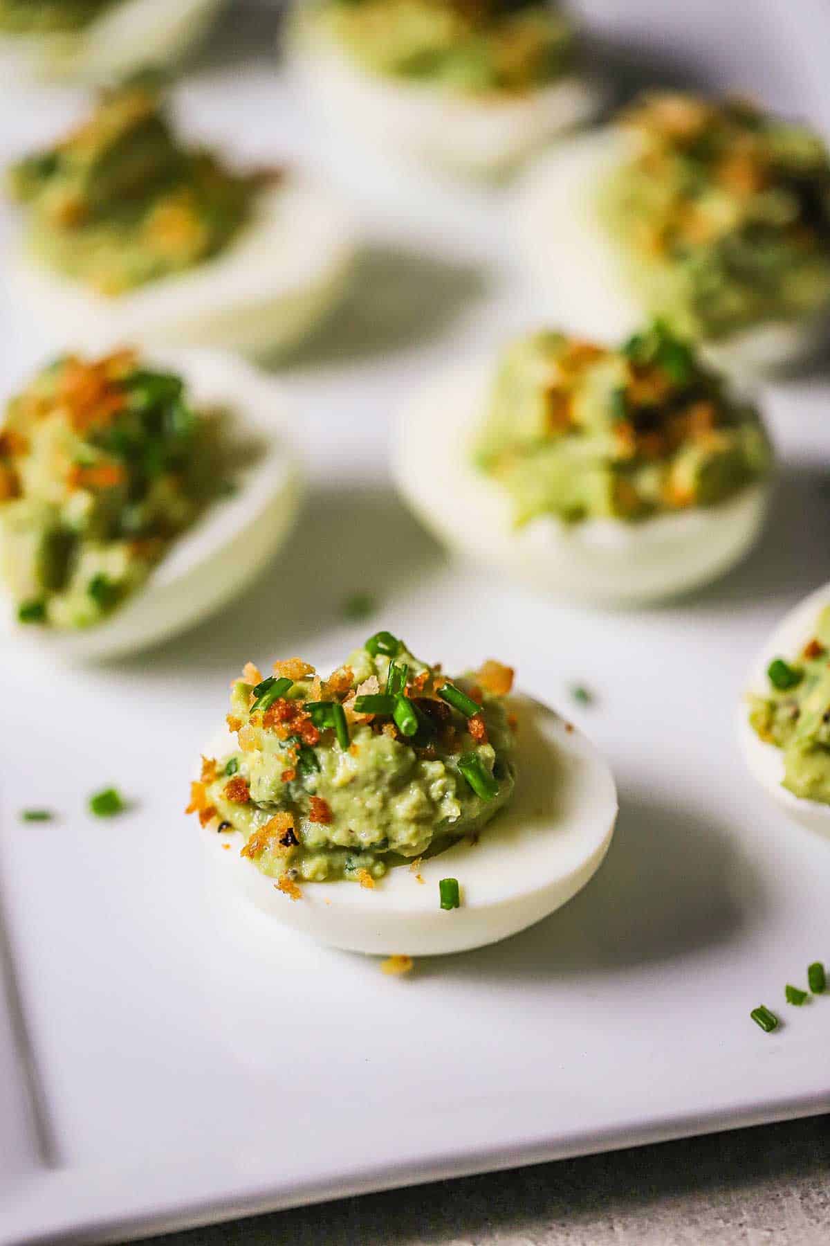 Several rows of avocado deviled eggs resting on a white platter and are garnished with toasted breadcrumbs, bacon, and chives.