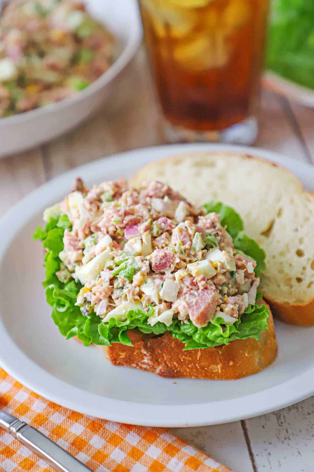A white plate topped with a thick slice of white bread topped with a layer of green leaf lettuce and a serving of ham salad.