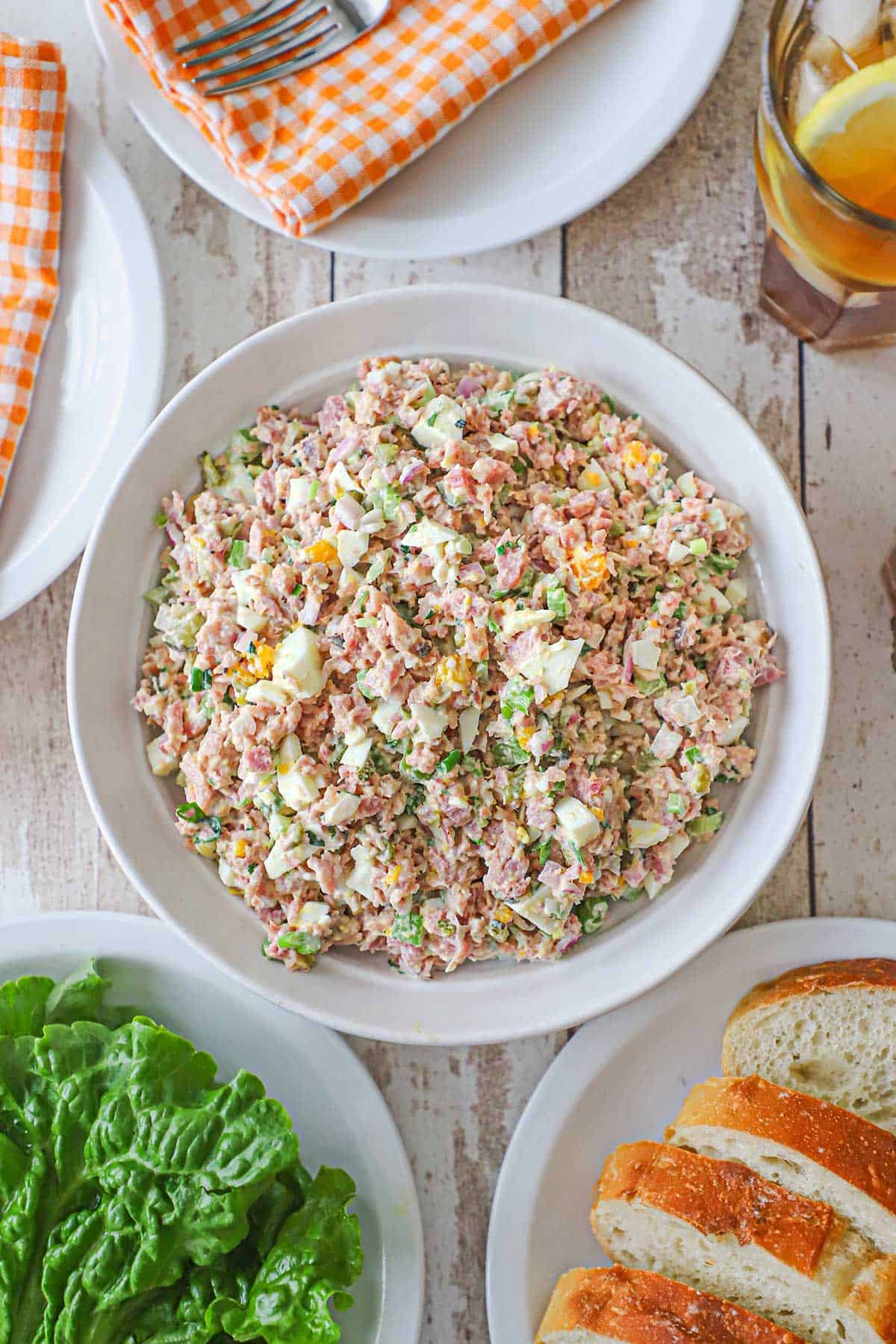 A shallow white bowl filled with best-ever ham salad surrounded by a plate of green leaf lettuces, slices of bread, plates, and glasses of iced tea.