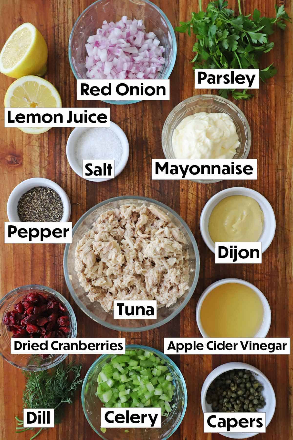 The ingredients for gourmet tuna salad arranged on a wooden surface, including a can of tuna, capers, fresh dill, dried cranberries, mayonnaise, Dijon mustard, white wine vinegar, and a selection of seasonings, all neatly organized for easy preparation.