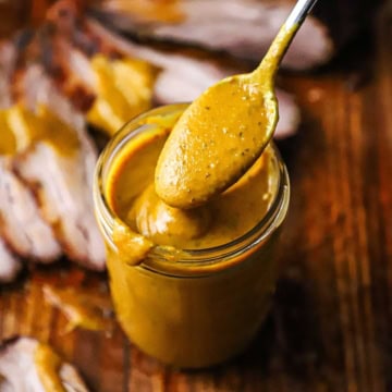 A spoon filled with Carolina mustard sauce being raised from a jar filled with the sauce with a sliced brisket nearby.