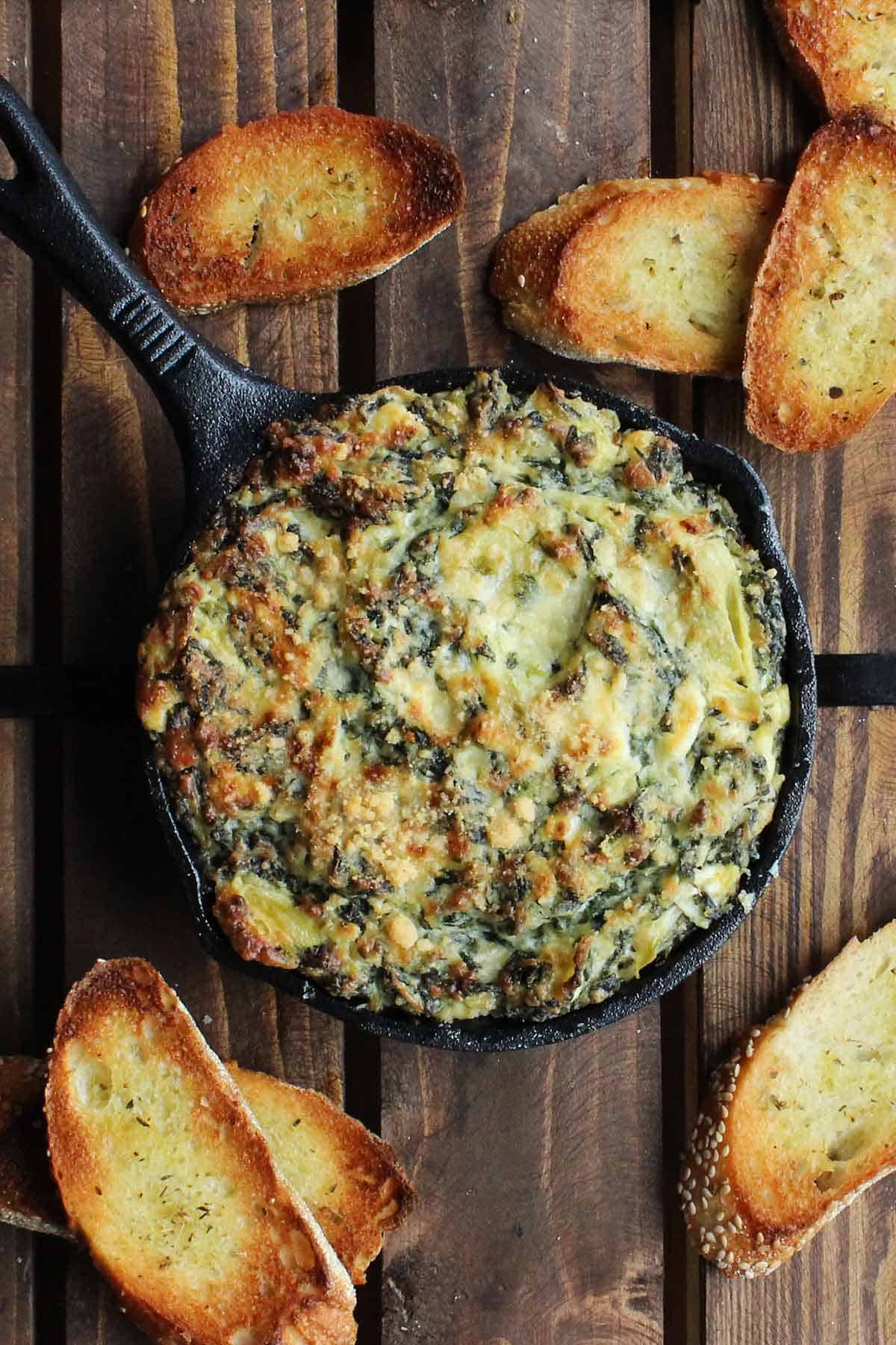 A small cast-iron skillet filled with baked spinach and artichoke dip with several small slices of toasted baguette nearby.