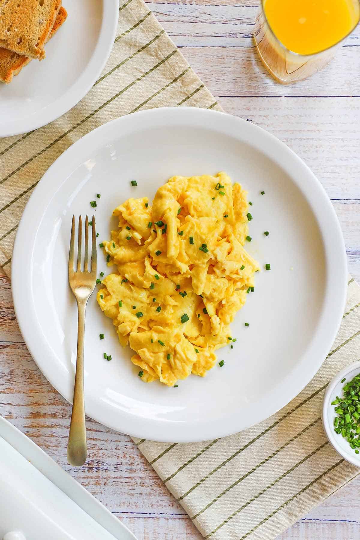 A white plate filled with the fluffiest scrambled eggs that have been garnished with snipped chives and a glass of orange juice nearby. 