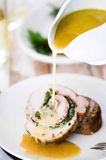 A person pouring brown gravy from a gravy boat onto a plate holding two slices of herb-stuffed pork loin.