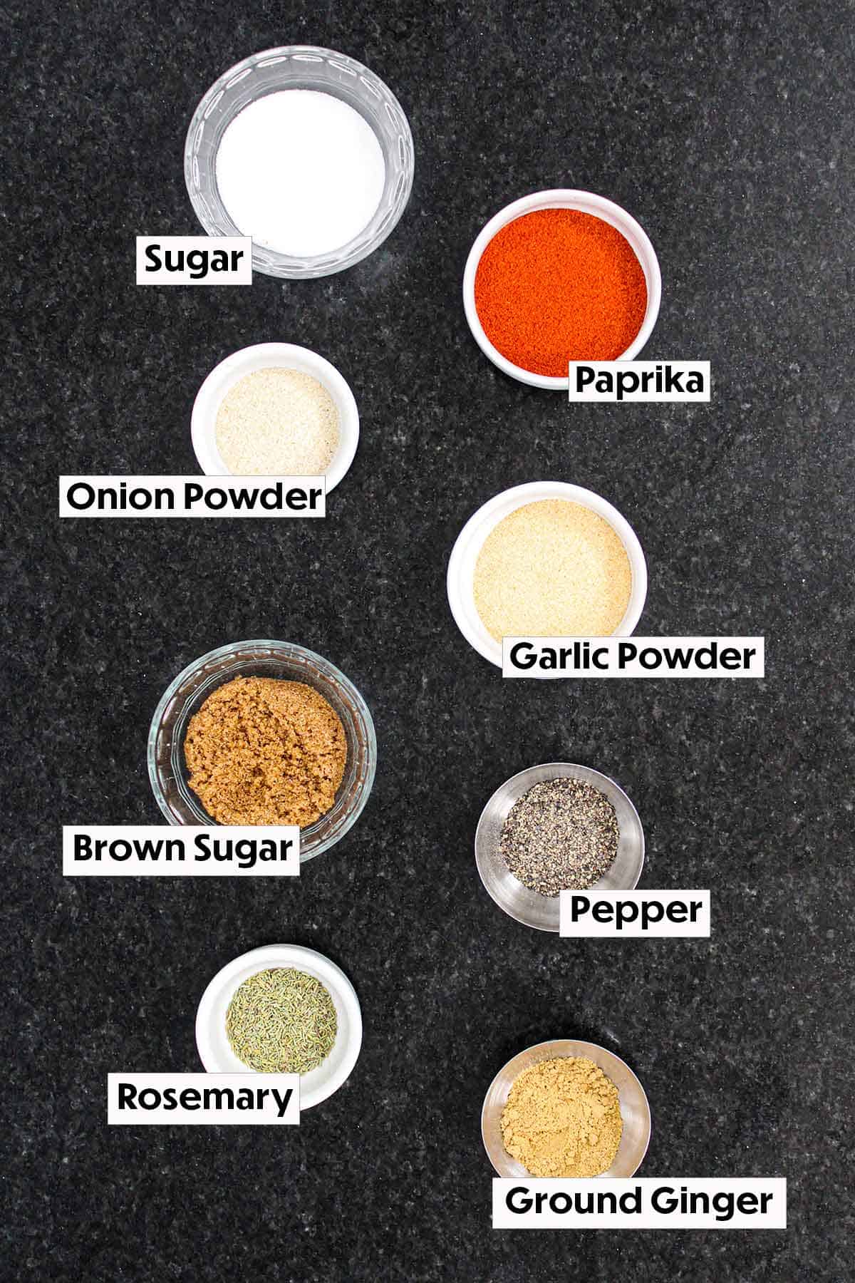 The ingredients for pork rub arranged on a black marble surface including sugar, paprika, onion and garlic powders, brown sugar, and seasonings.