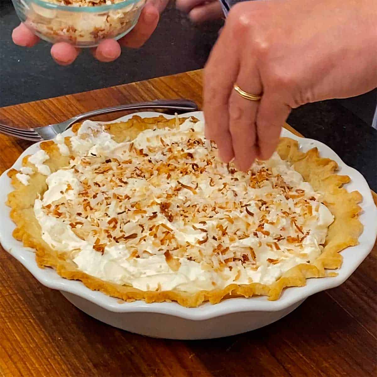 A person sprinkling toasted coconut flakes over the top of a coconut cream pie.