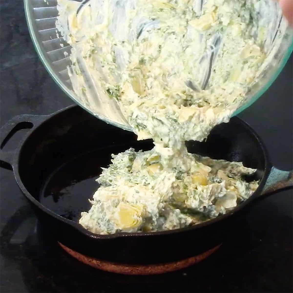 A person transferring uncooked spinach and artichoke dip into a small cast-iorn skillet.