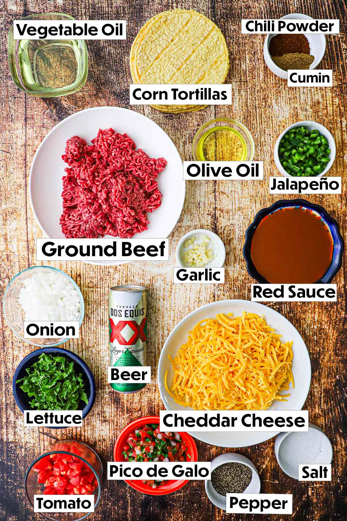 The ingredients for red enchilada sauce arranged on a wooden background, including ground beef, shredded cheese, corn tortillas, beer, garlic cloves, chopped onion, and a variety of spices, all neatly organized for easy preparation.