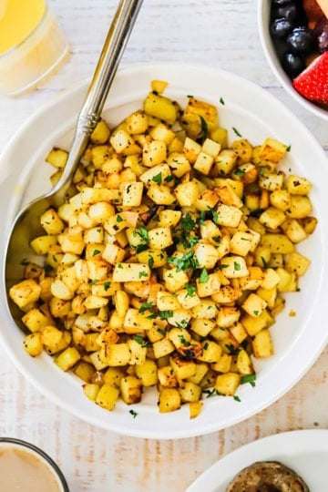 A white bowl filled with easy breakfast potatoes garnished with chopped parsley with a large serving spoon inserted along the side of the dish.