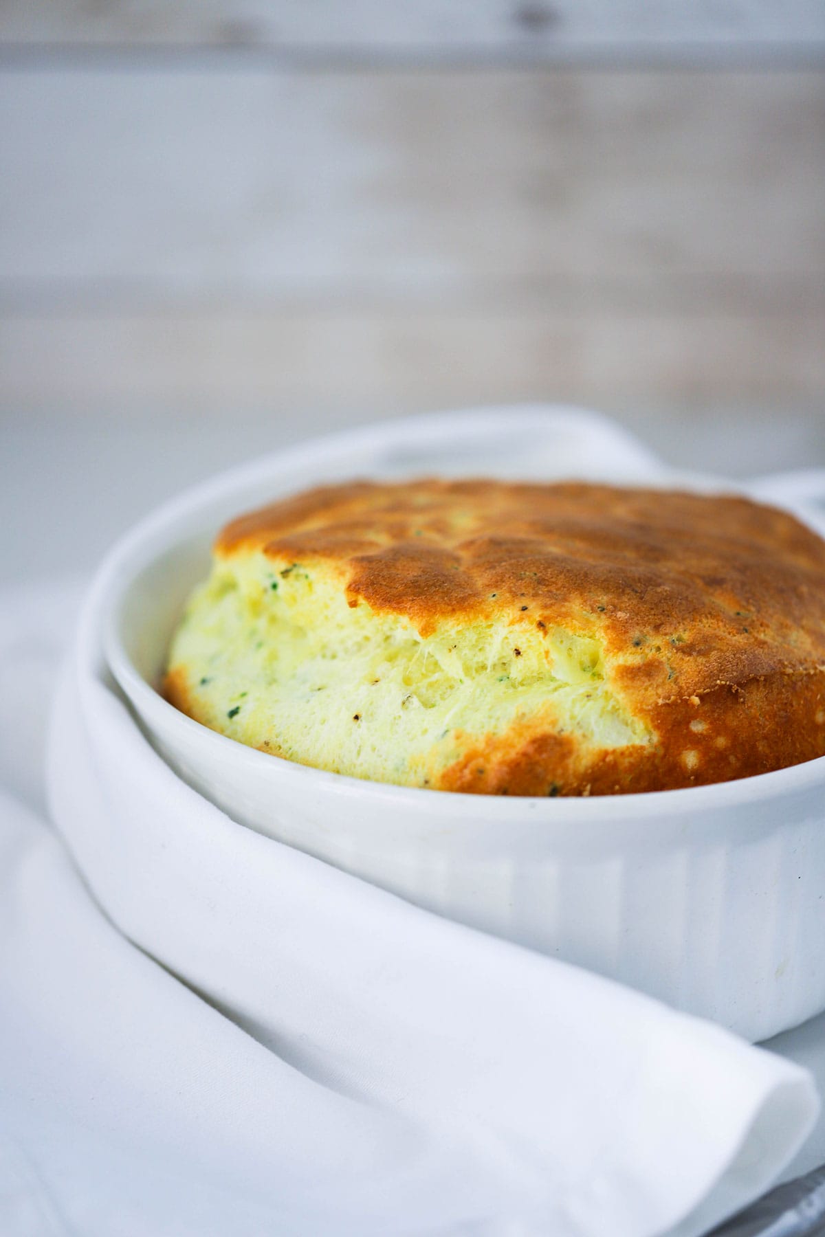 A fully cooked cheese soufflé with cheese in a white dish surrounded by a white linen napkin.