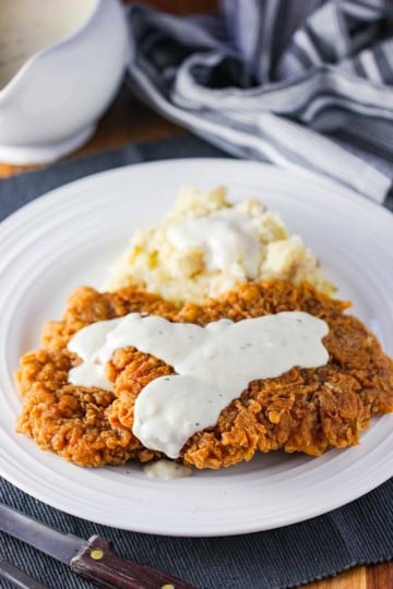A crispy Southern chicken fried steak on a white dinner plate topped with cream gravy and next to a pile of mashed potatoes.