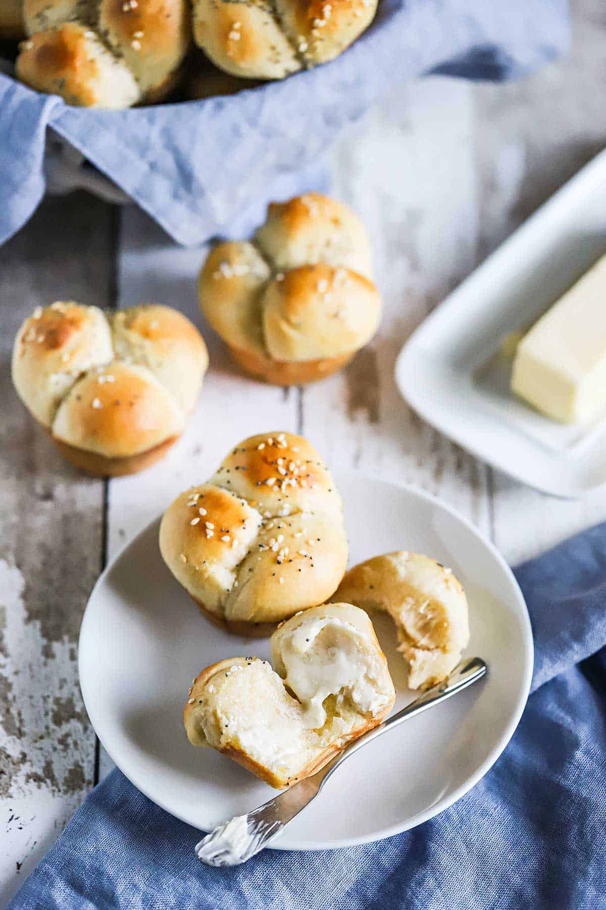 Several cloverleaf dinner rolls arrange on a white background with one of them on a small plate and is broken open with softened butter smeared on it.