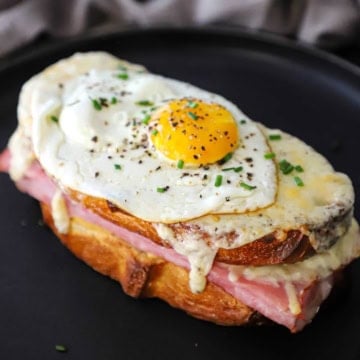 A Croque Madame sandwich with a sunny side egg resting on top of it on a black plate.