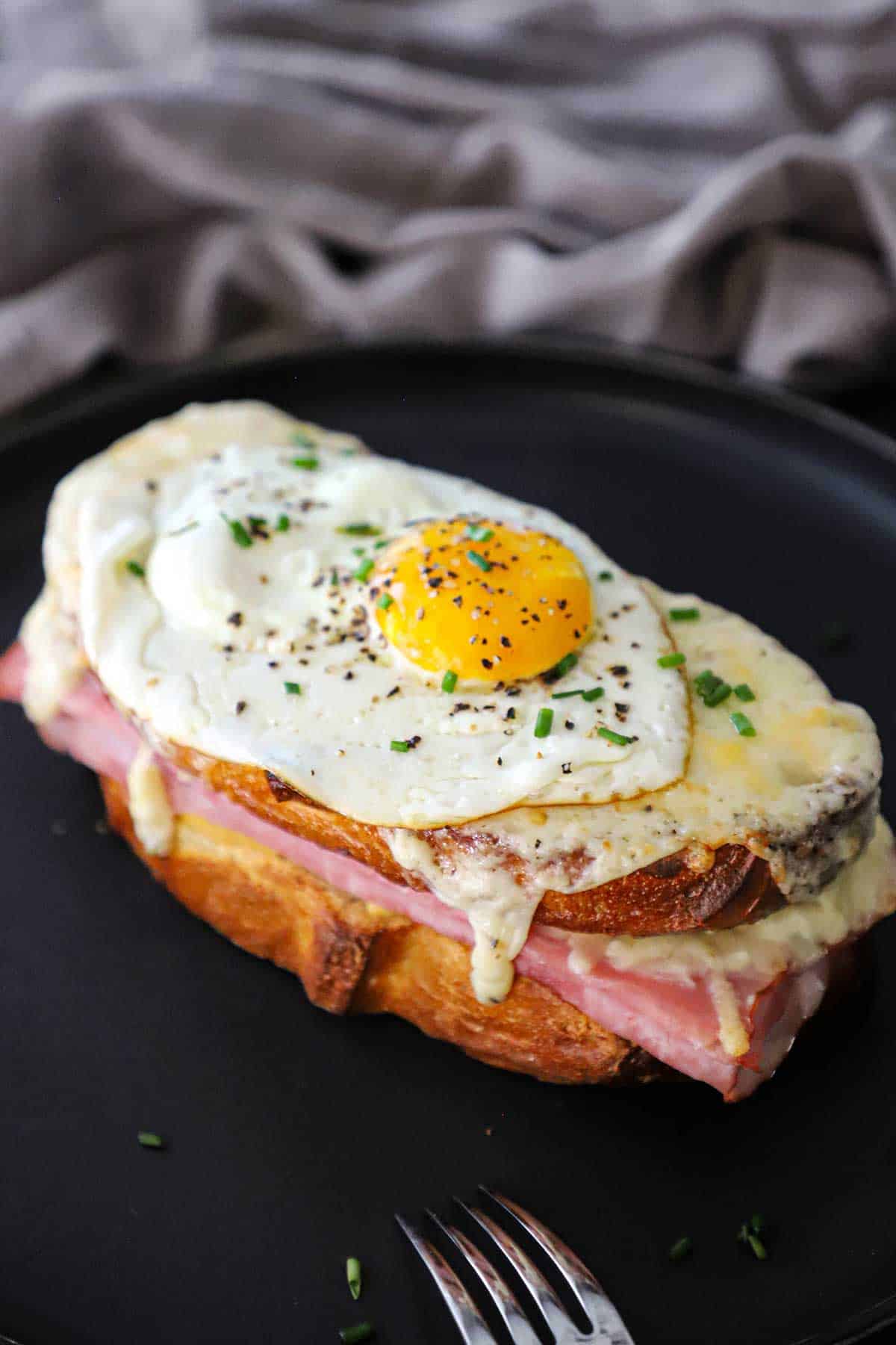 A Croque Madame sandwich with a sunny side egg resting on top of it on a black plate.