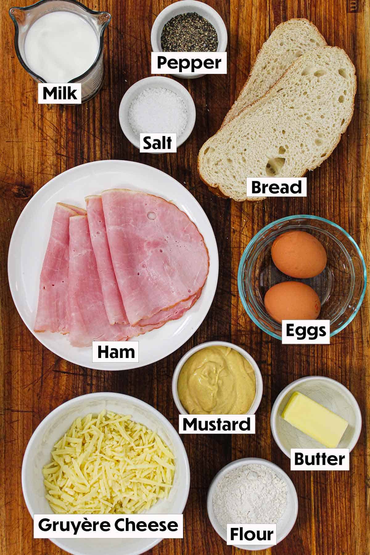 The ingredients for a Croque Madame arranged on a wooden background, including slices of bread, ham, Gruyère cheese, eggs, and a small bowl of homemade mornay sauce, all neatly organized for easy preparation.